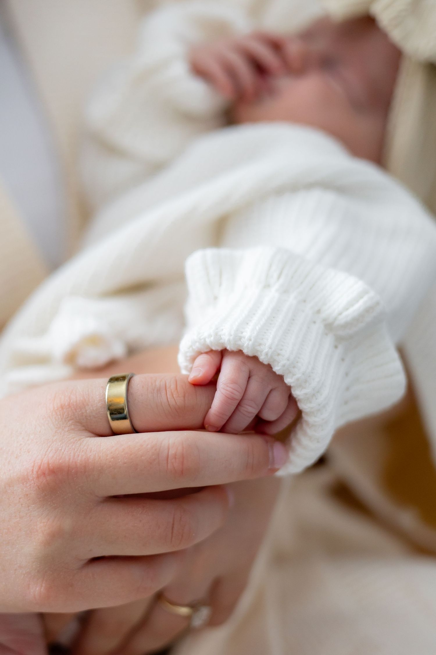 A close-up of a tiny baby hand holding an adult's finger with wedding ring visible.