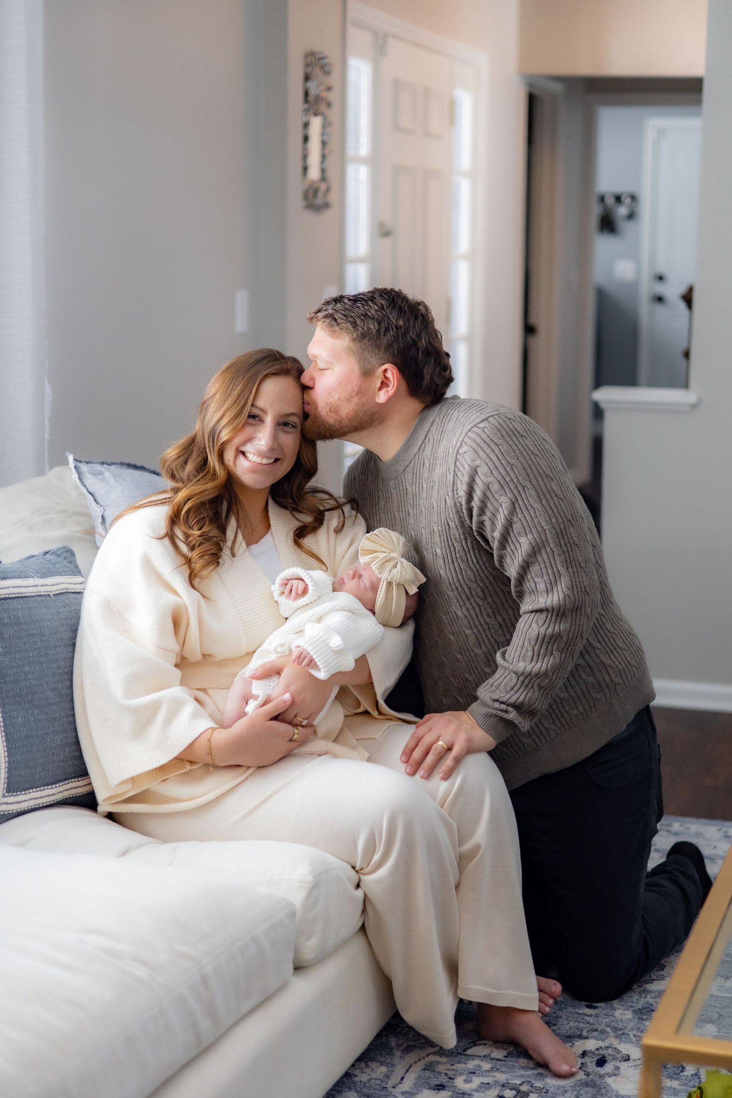 A couple sits together on a white couch sharing tender moments with their newborn baby in a bright home setting.