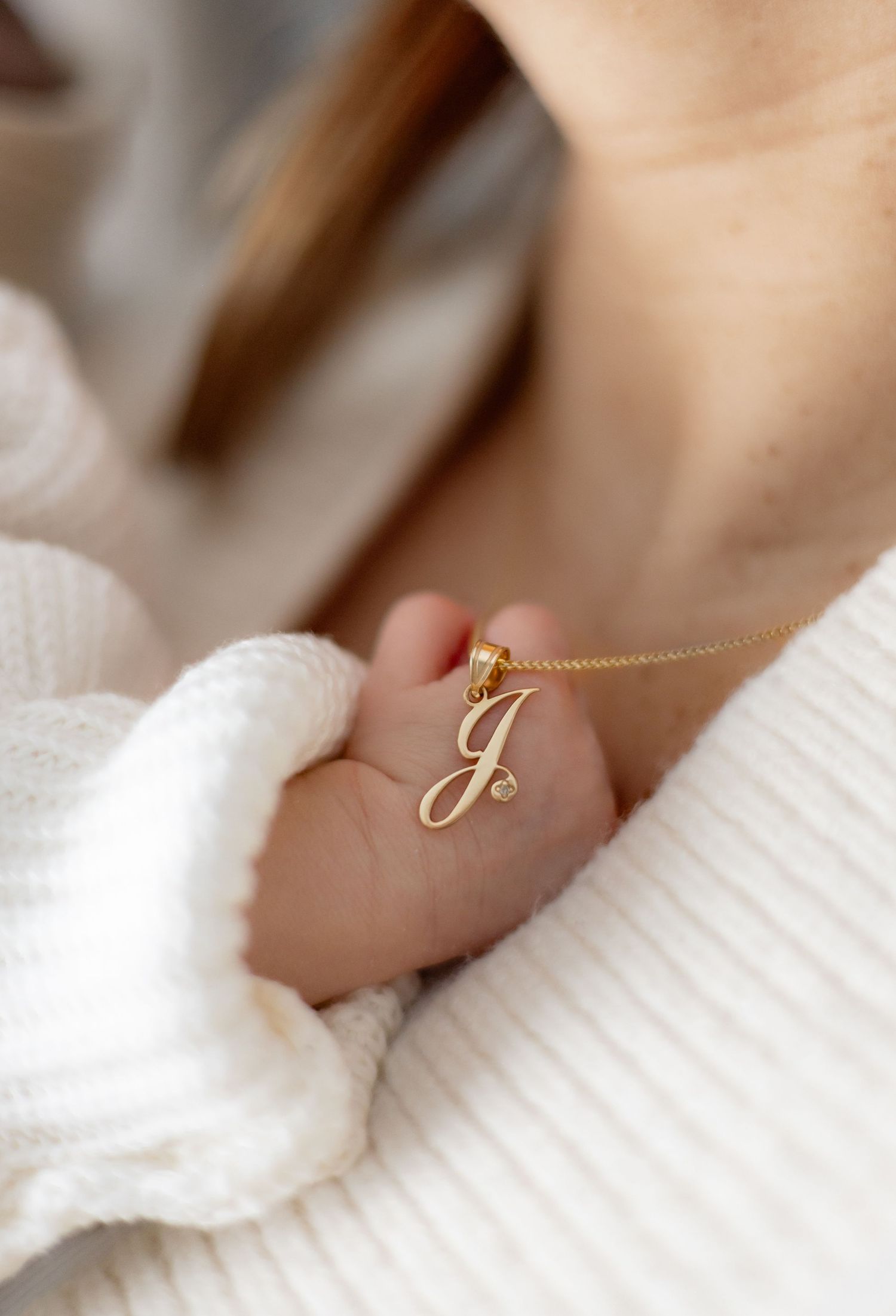 A delicate gold initial necklace rests against soft white fabric with a baby's hand visible.