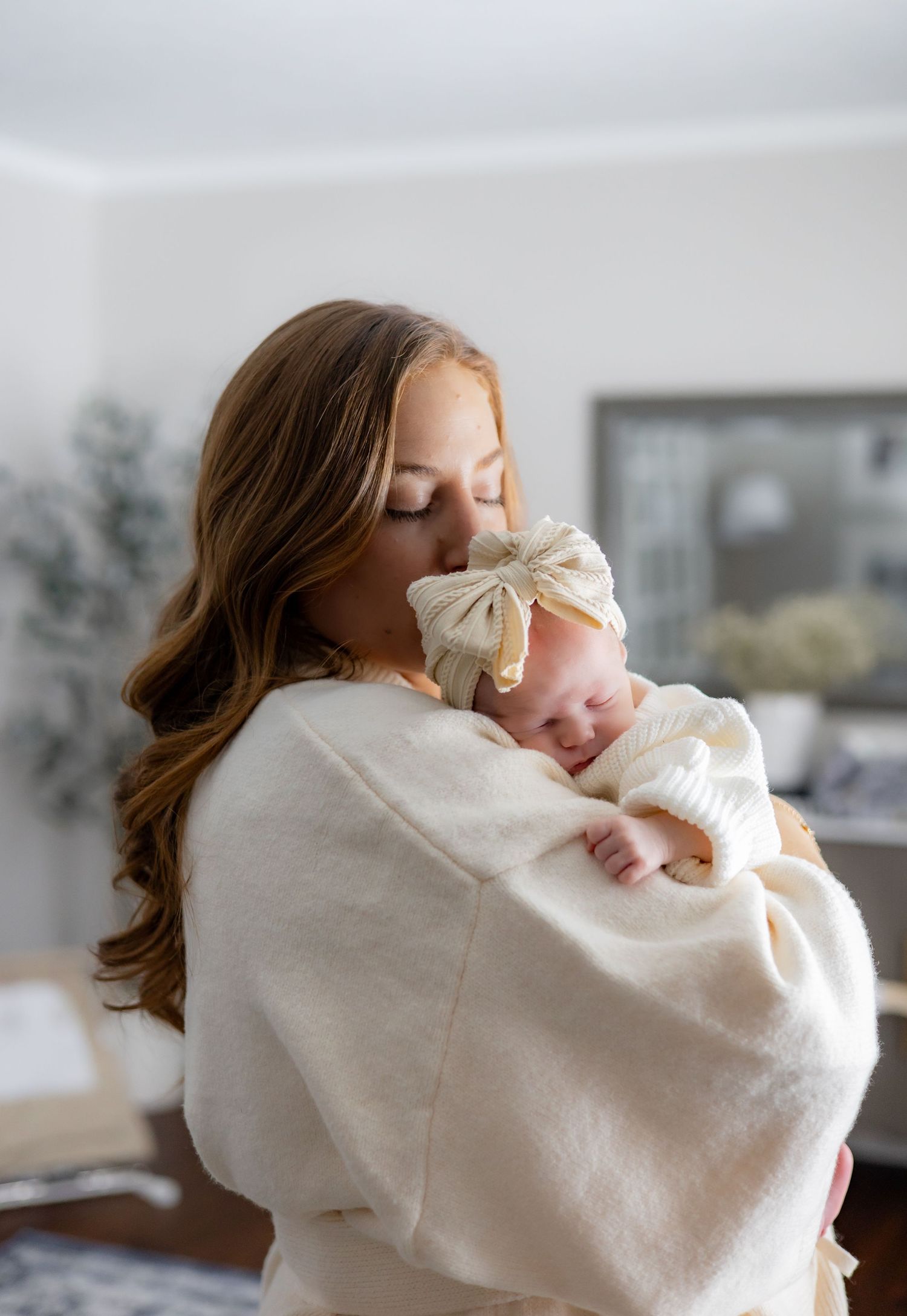 Person in cream sweater holds and cradles small baby in bright room.