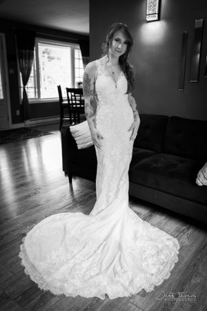 A bride in a flowing lace gown with intricate tattoos stands elegantly in a cozy living room. Jeff Fevens Photography in Fredericton NB.