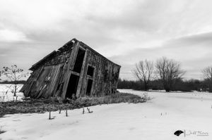 A weathered, leaning barn stands alone in a snowy field under a moody sky, evoking time’s passage. Jeff Fevens Photography in Fredericton NB