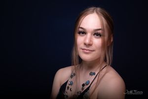 A young woman with long brown hair and a gemstone necklace poses against a deep blue backdrop. Jeff Fevens Photography in Fredericton