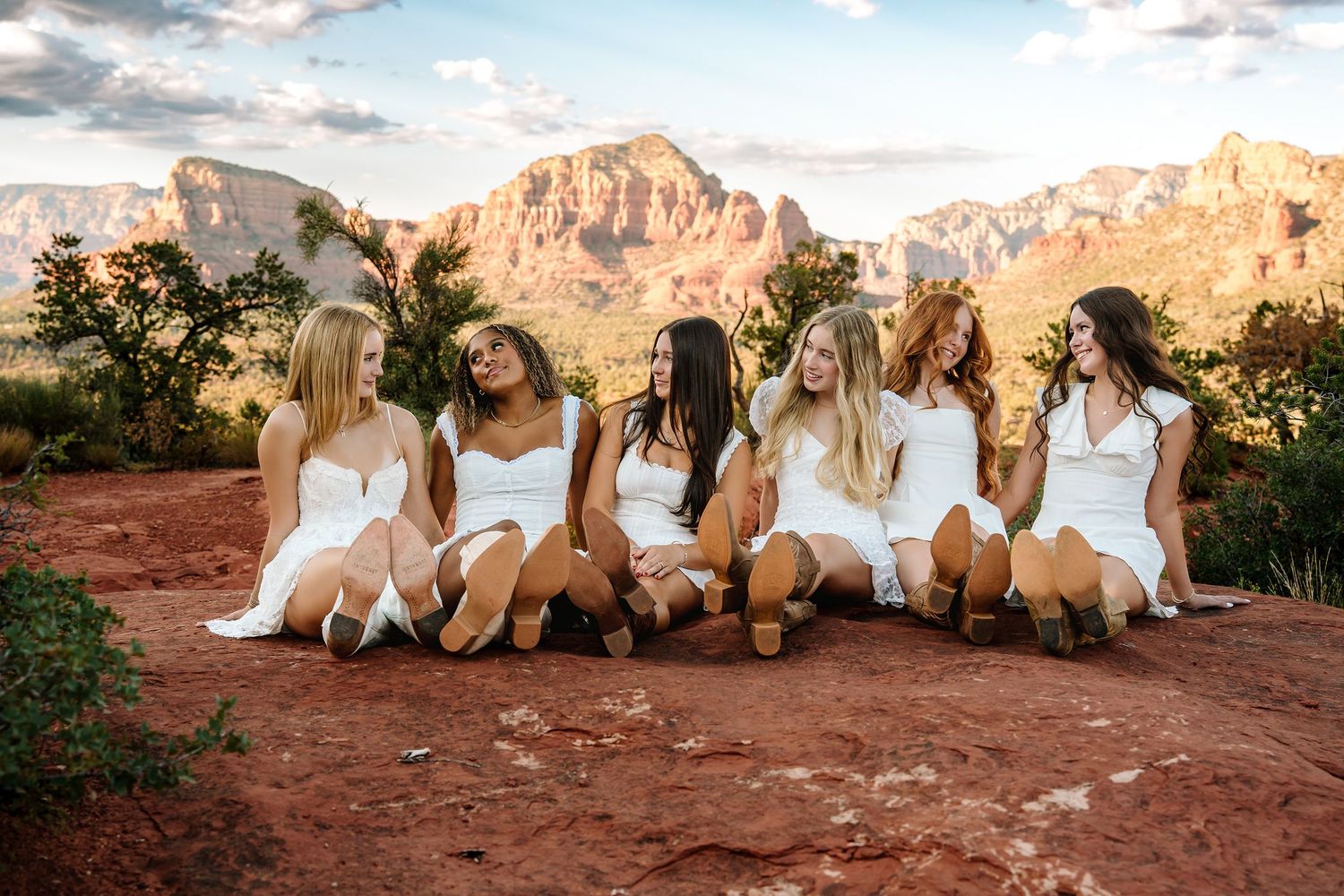 Six high school seniors in white dresses sit together on red rock in Sedona, Arizona, with stunning red rock formations in the background.
