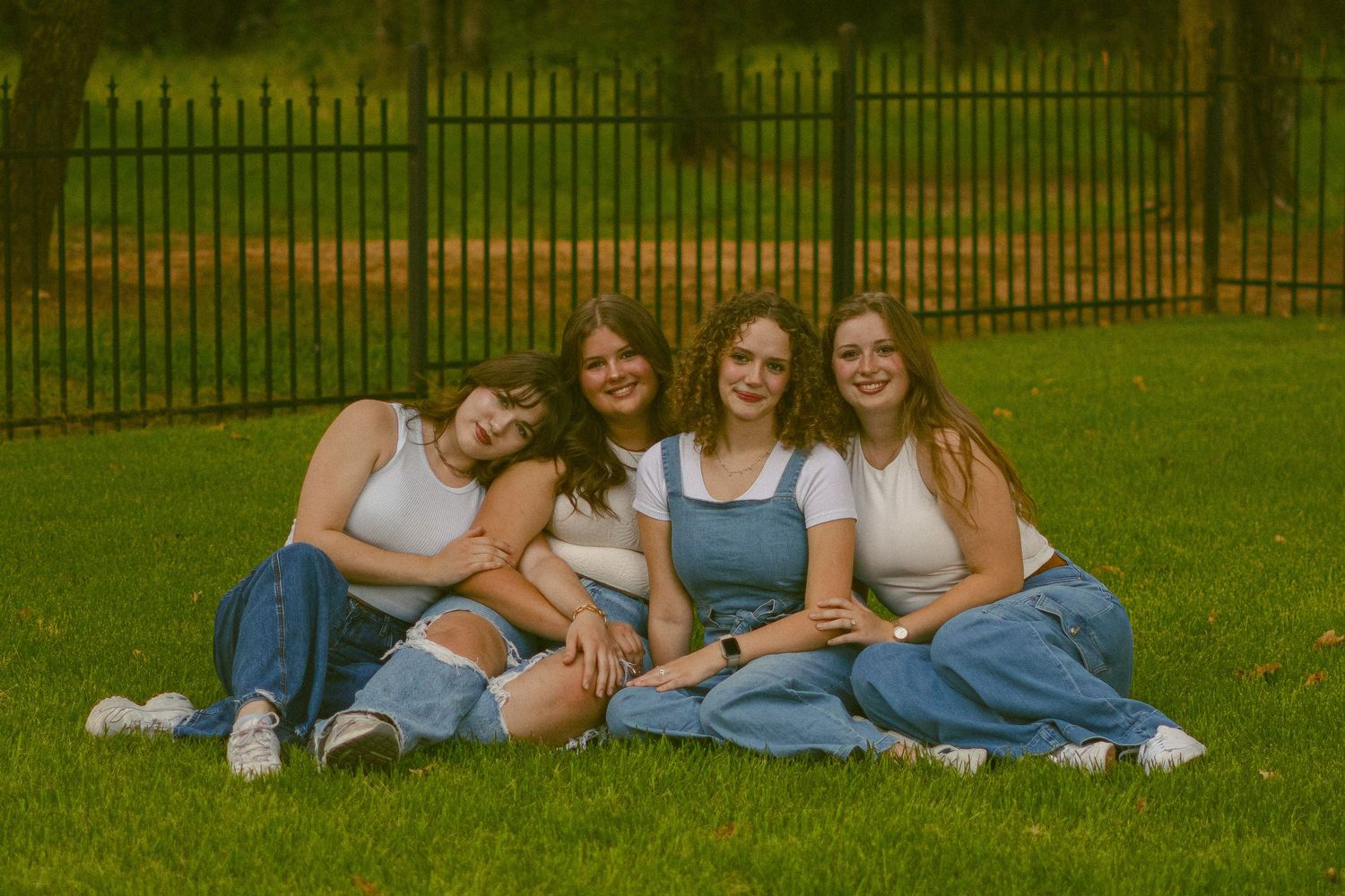 Friends in coordinated denim outfits relax on lawn near iron fence at sunset.