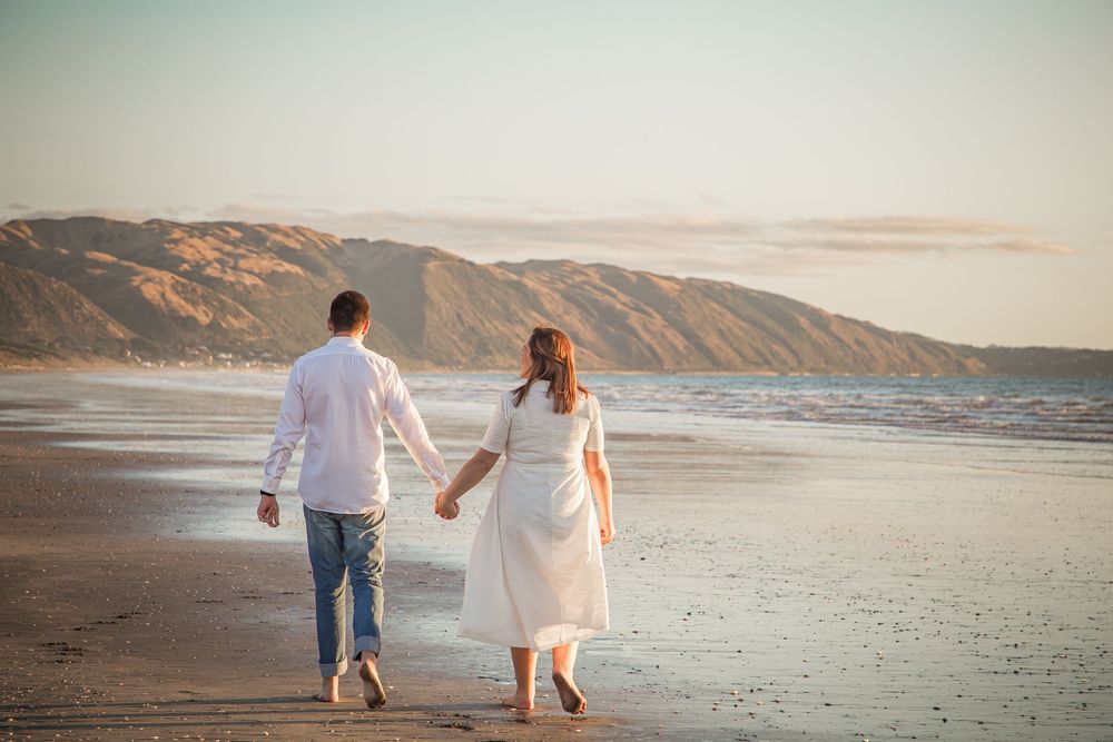 Romantic couple walking hand in hand along a scenic beach at sunset with mountains in the background.