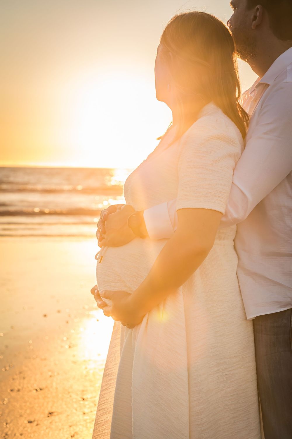 A couple embraces on a beach at sunset, silhouetted against the golden light reflecting off the ocean waves.