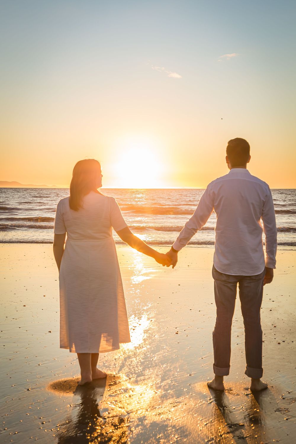 Silhouetted couple holding hands while walking along a beach at sunset with golden light reflecting on wet sand.