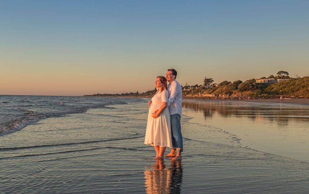 Couple standing together on beach at sunset with waves lapping at their feet and coastal homes in background.