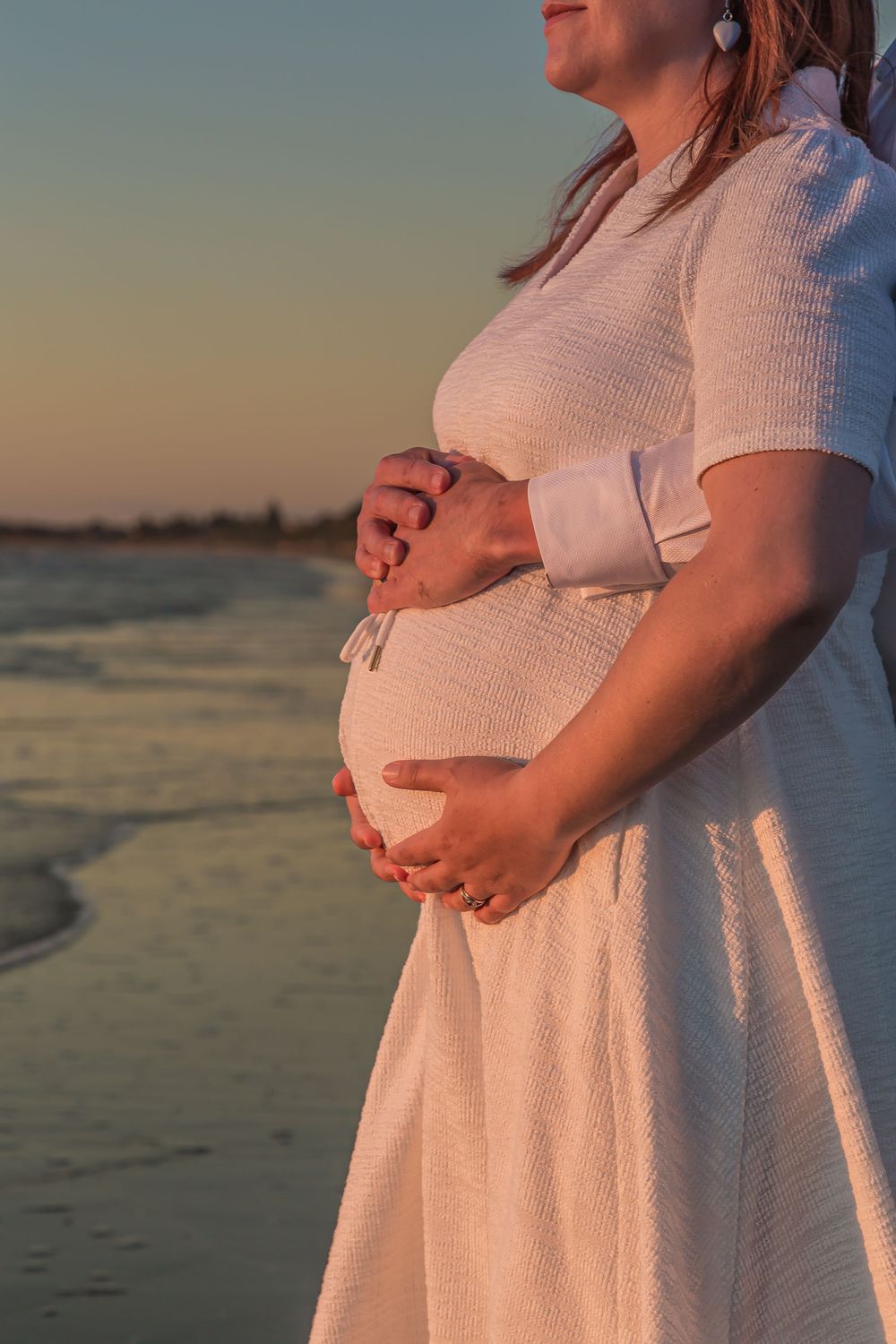 A pregnant woman in a white dress stands on the beach at sunset, cradling her baby bump in soft golden light.