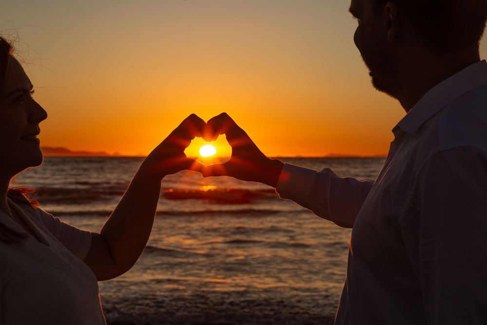 Silhouetted figures hold up the sun during a vibrant orange sunset over the ocean waves.