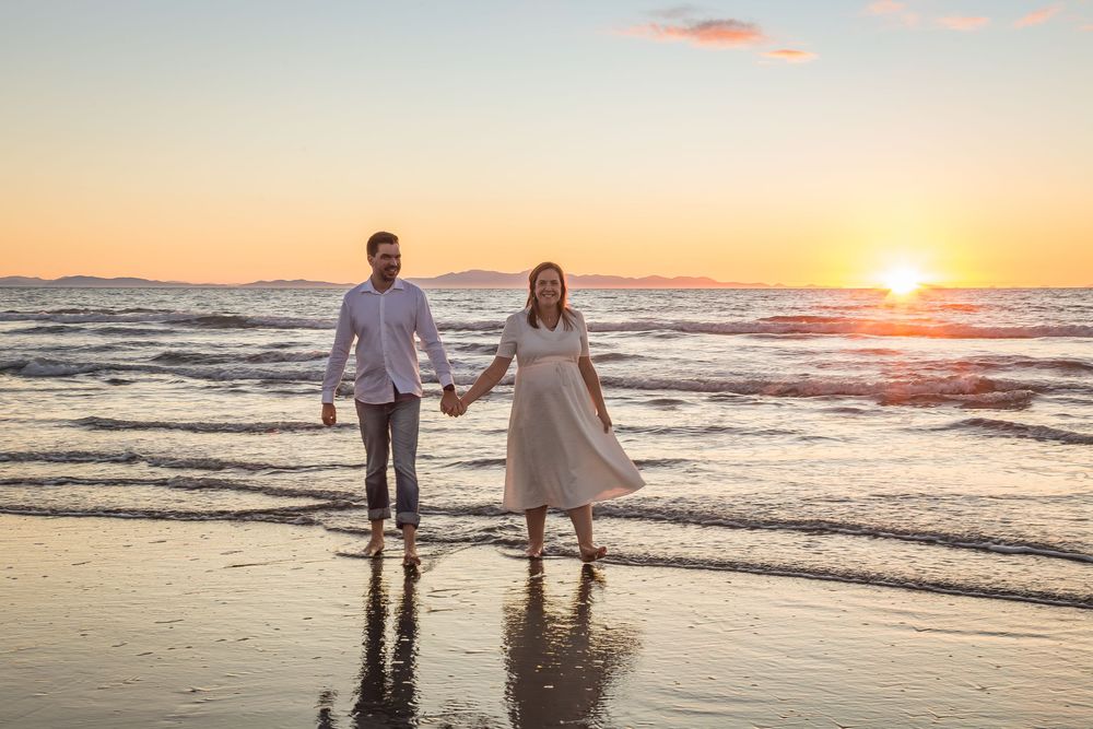 A couple holds hands while walking along a beach at sunset with waves crashing on the shore.