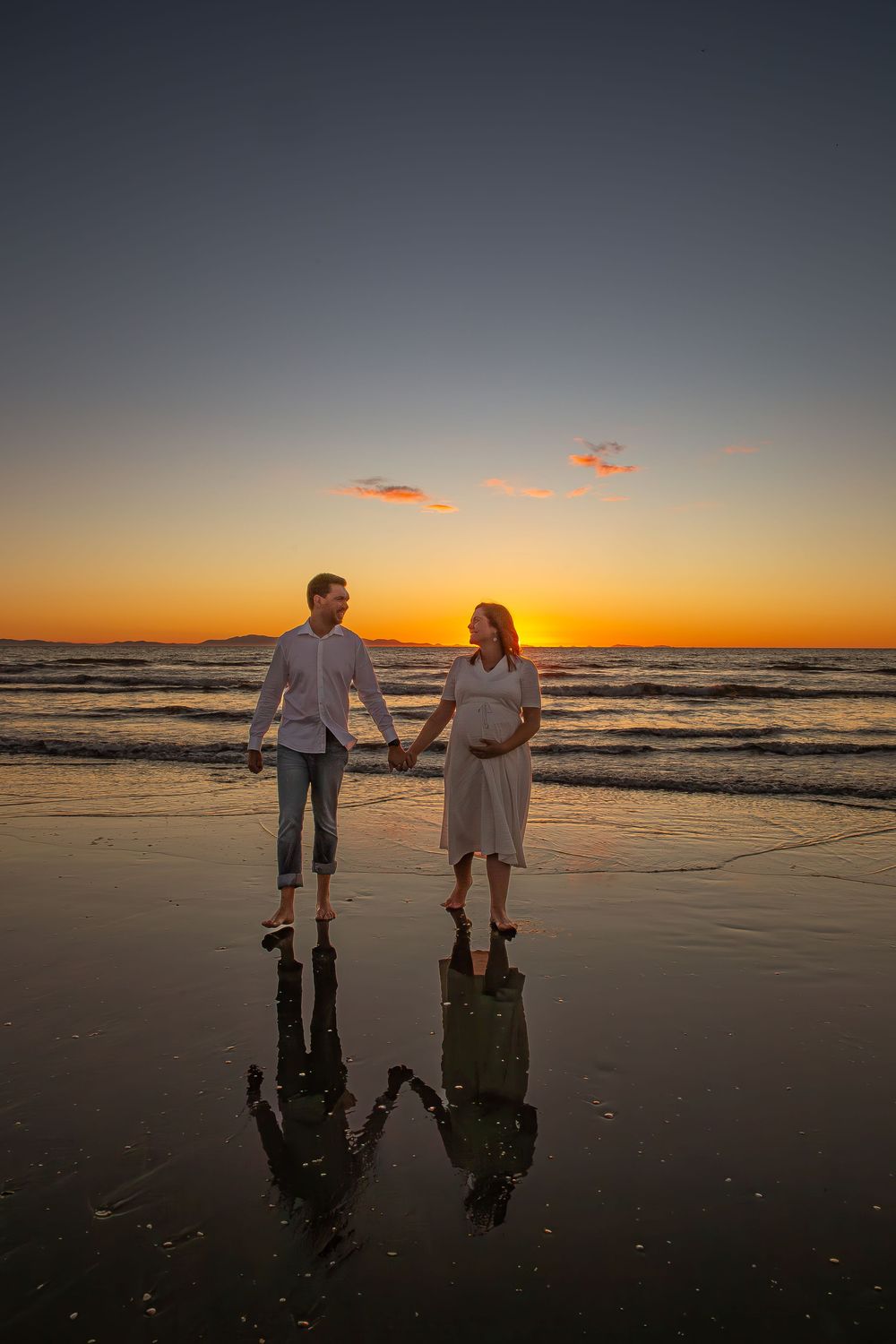 Two figures stand silhouetted against a vibrant orange sunset on a reflective beach shoreline with rolling waves.