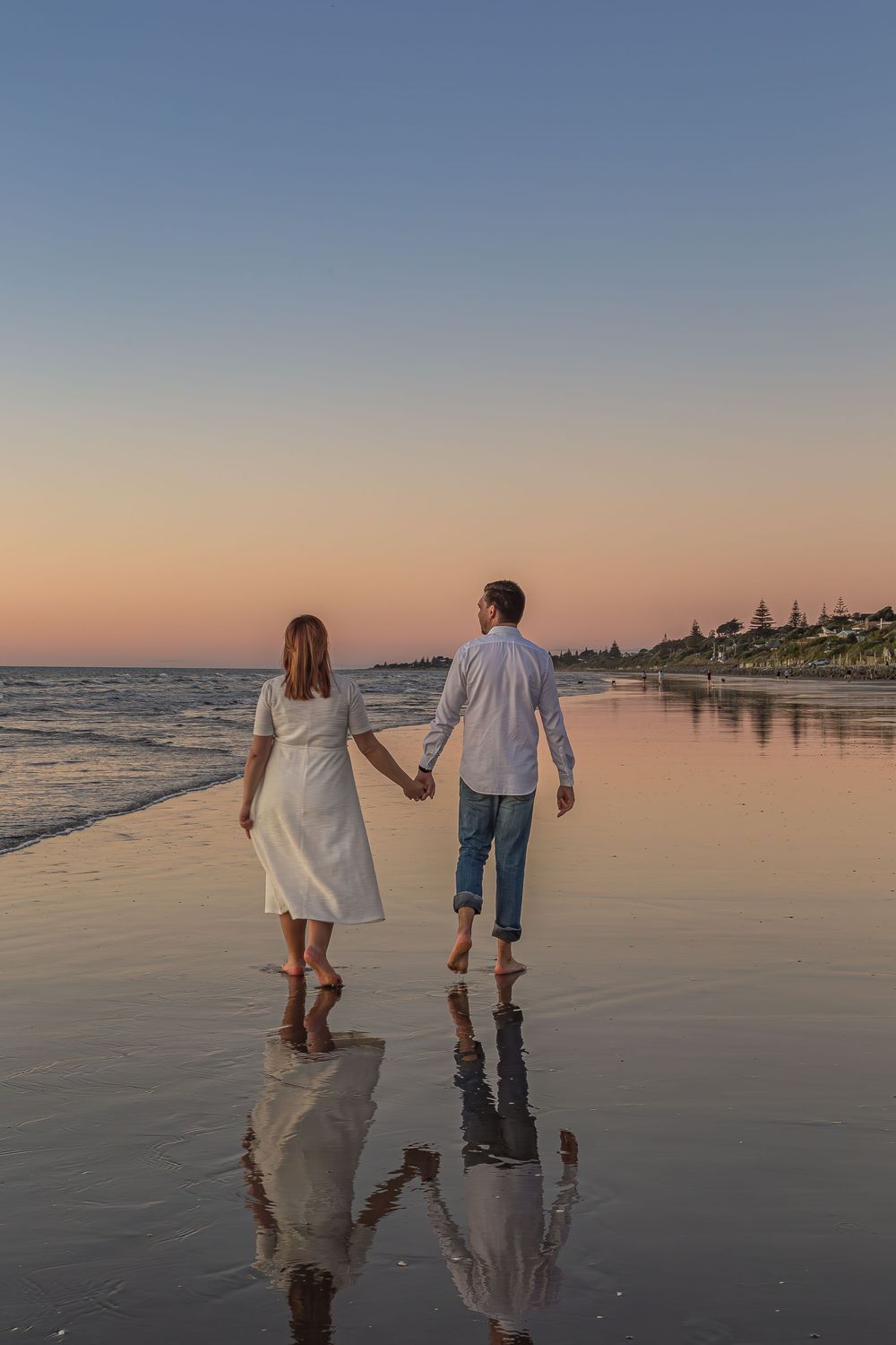 A couple walks hand in hand along a reflective beach shoreline during sunset, creating mirror-like silhouettes on wet sand.