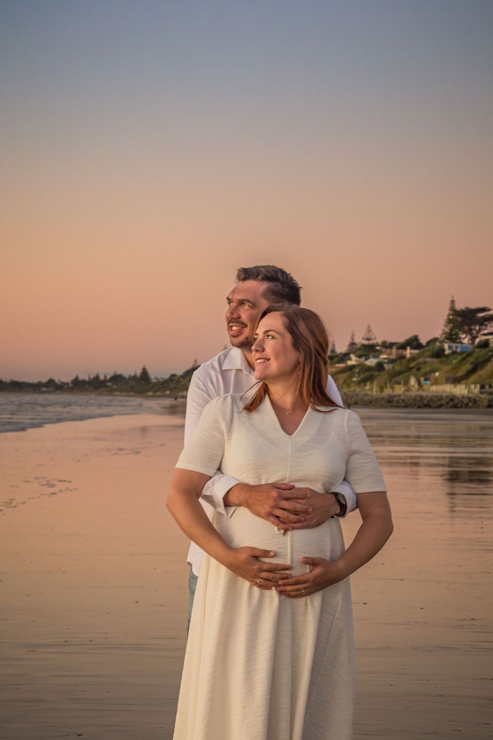 A couple in white outfits embraces on a serene beach during golden hour sunset.