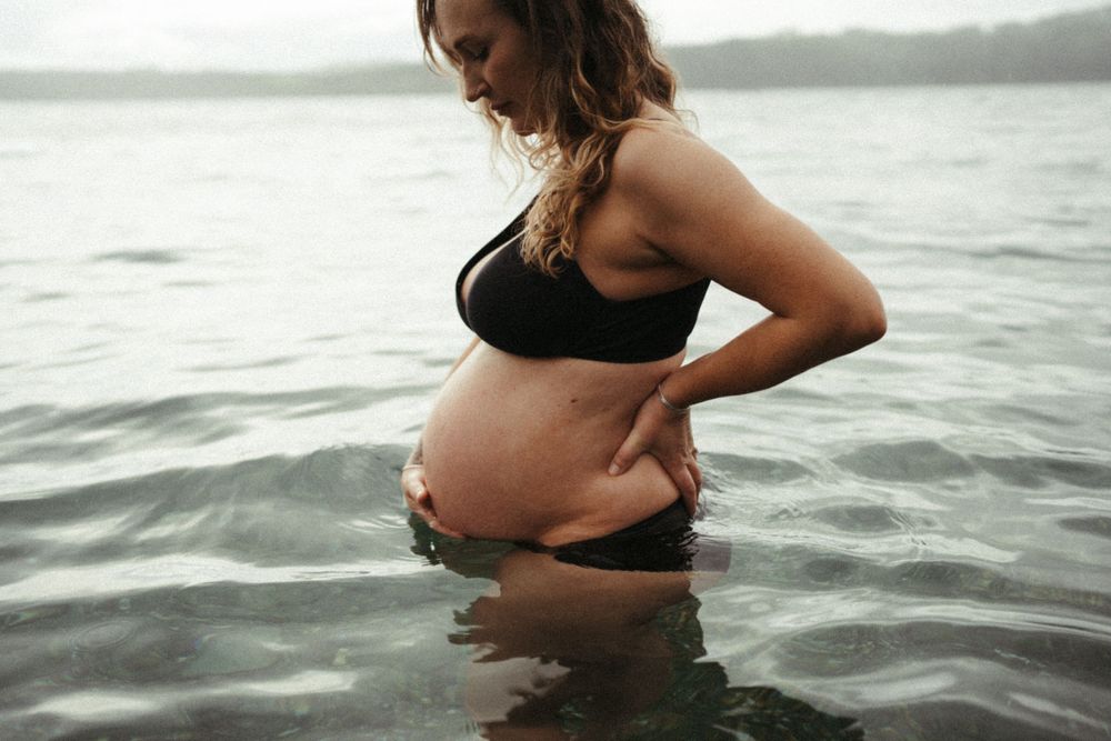 Pregnant woman in black bikini standing in ocean water touching her belly.