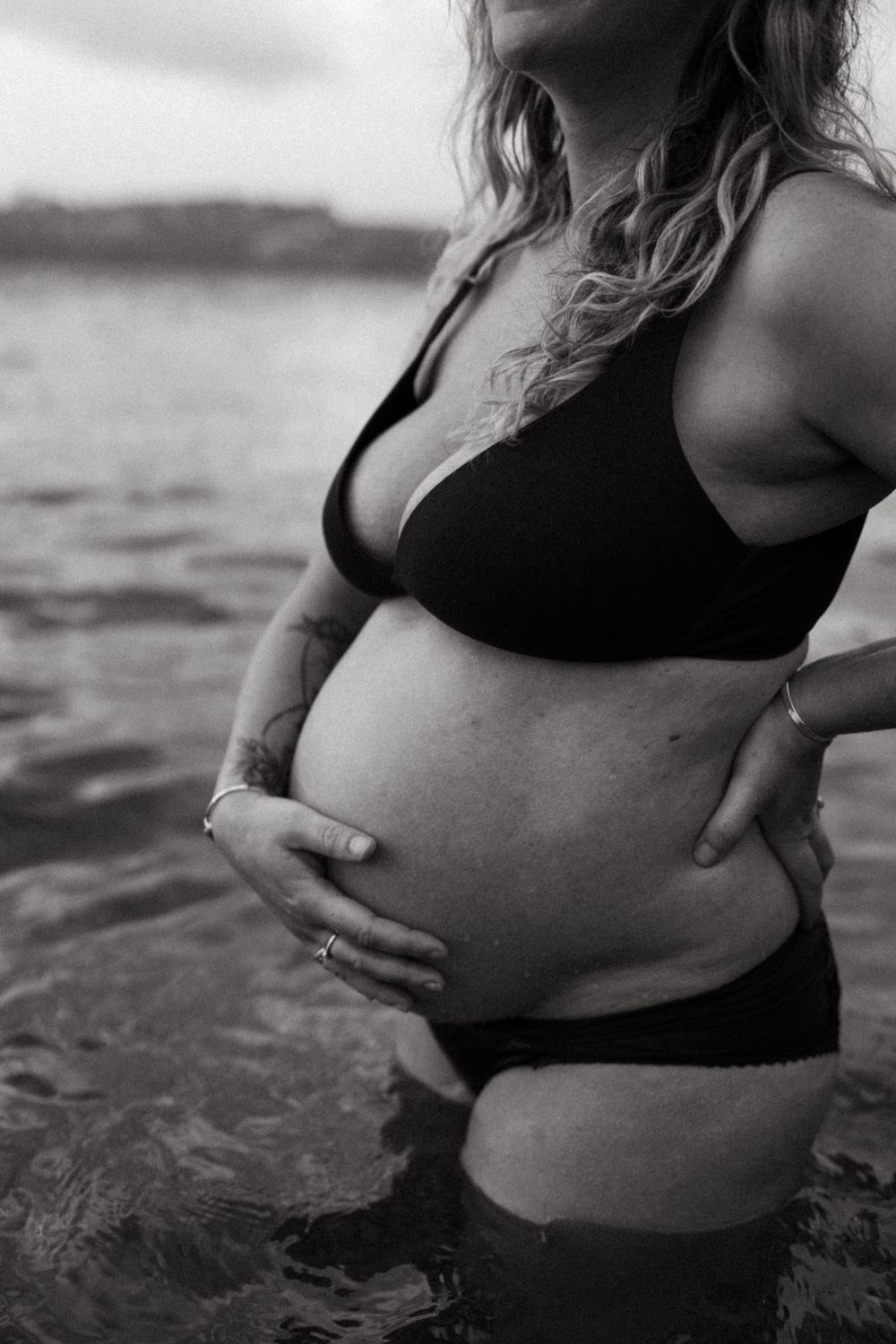 Black and white photo of pregnant woman in swimwear standing in ocean water.