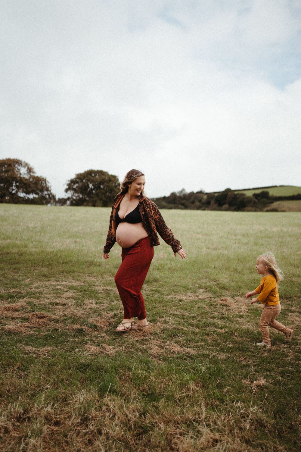 Pregnant woman in maroon pants plays in grassy field with small child.
