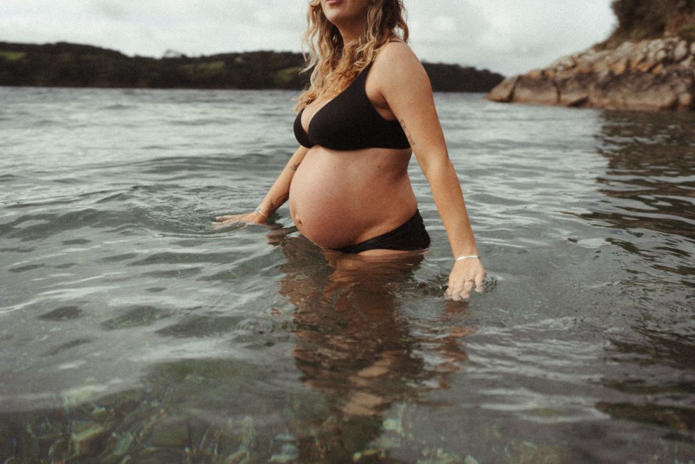 Expectant mother in black swimwear standing in shallow ocean water during sunset.