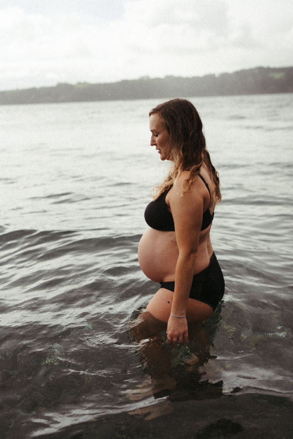 Pregnant woman in black swimsuit stands in calm ocean waters with cloudy sky.