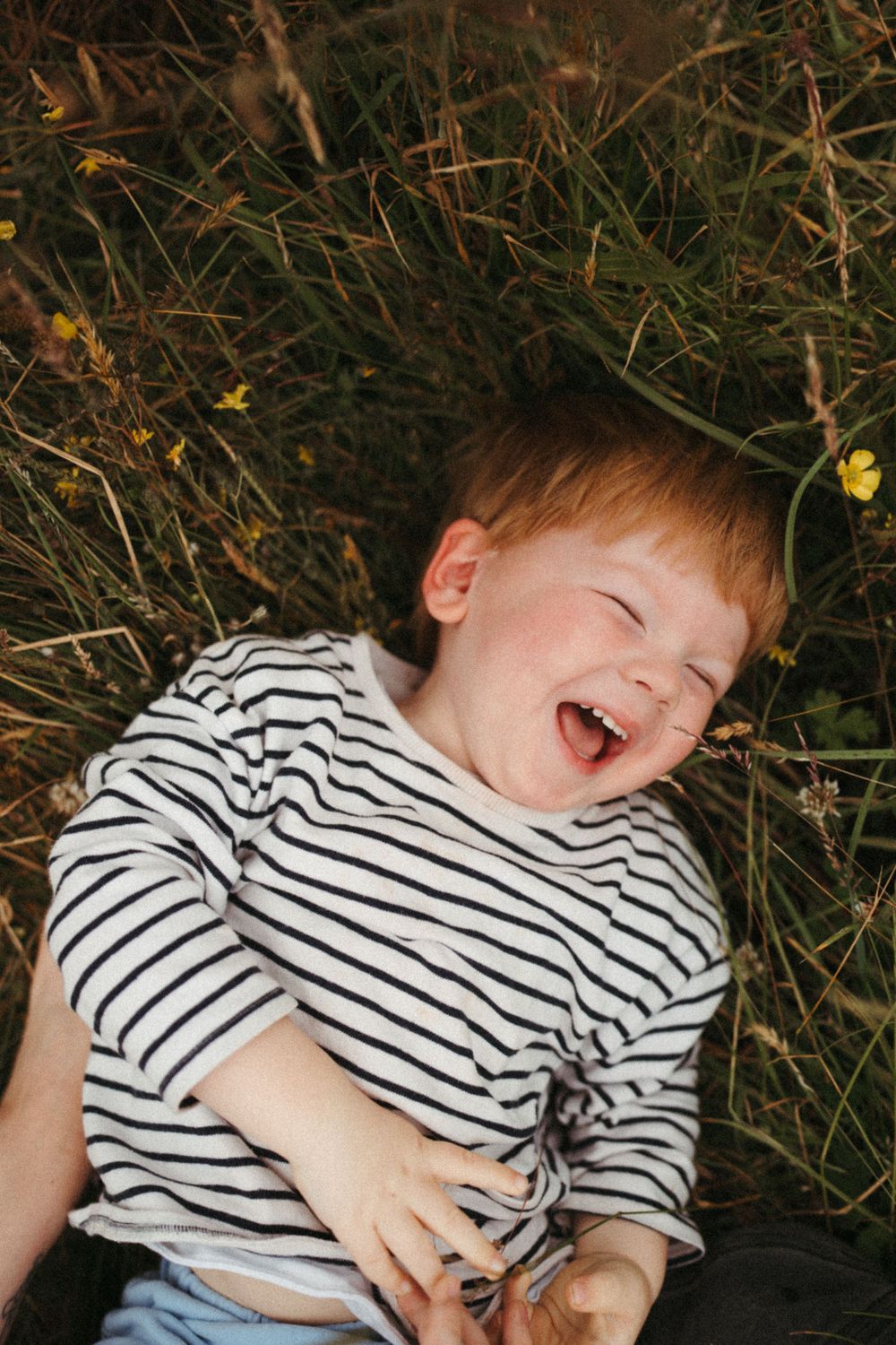 Child in striped shirt laughing joyfully while lying in tall grass with yellow flowers.