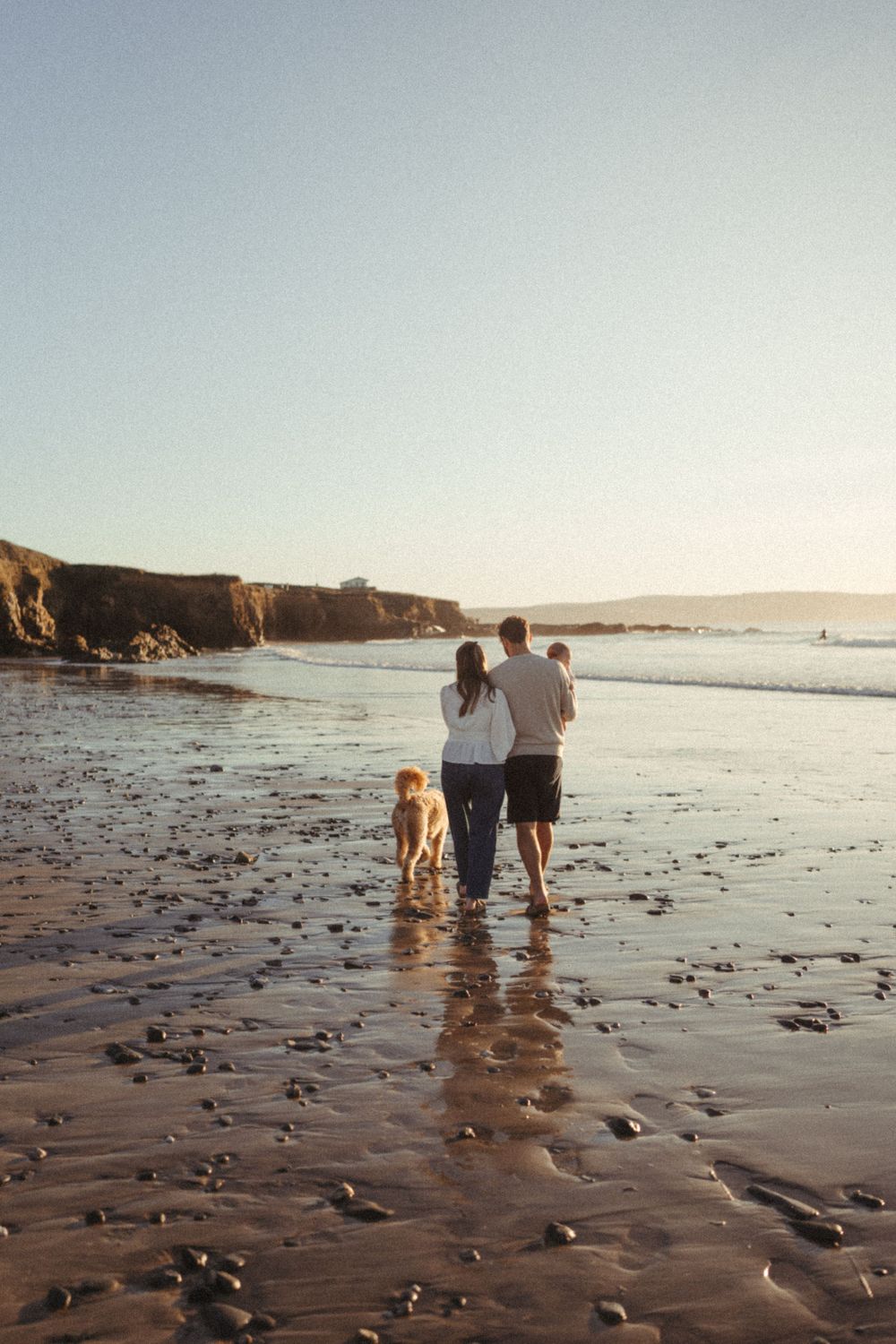 Couple walking with dog on scenic beach at sunset near rocky cliffs.