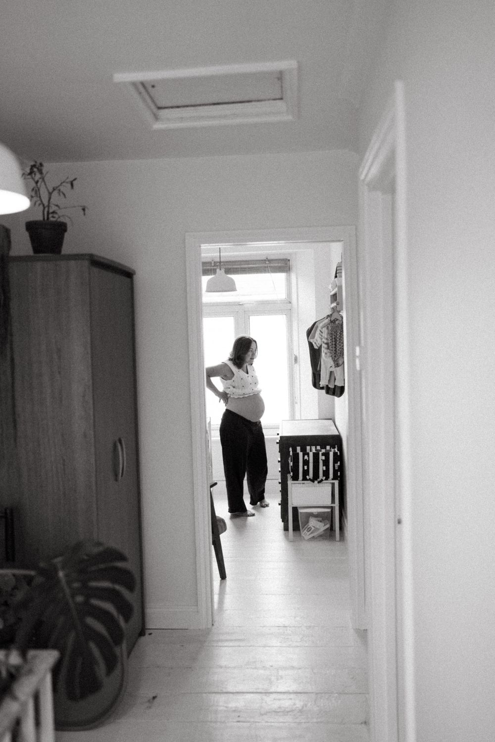 Black and white view down a hallway showing a figure in a doorway and wooden wardrobe against white walls.
