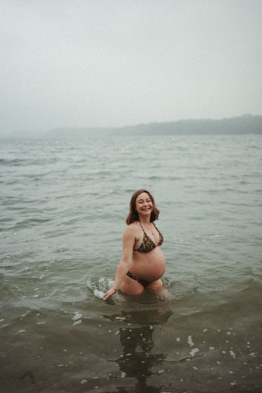A pregnant woman in a bikini stands in gray ocean waters on a misty day enjoying a refreshing swim.