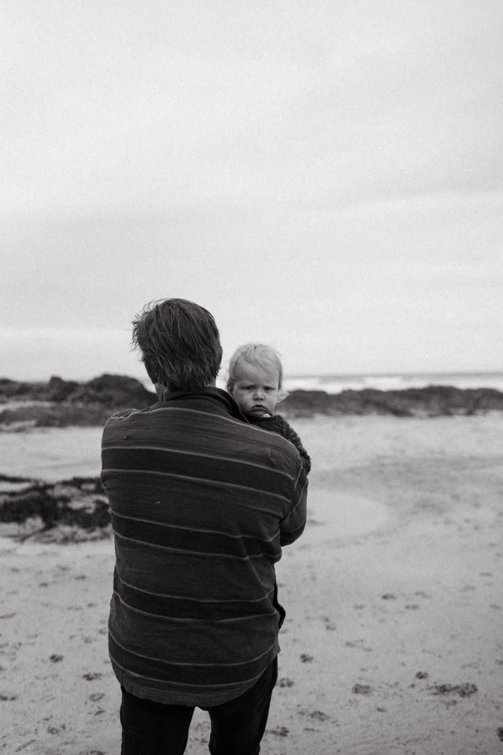 Black and white portrait of parent and child standing on a beach looking at the ocean.