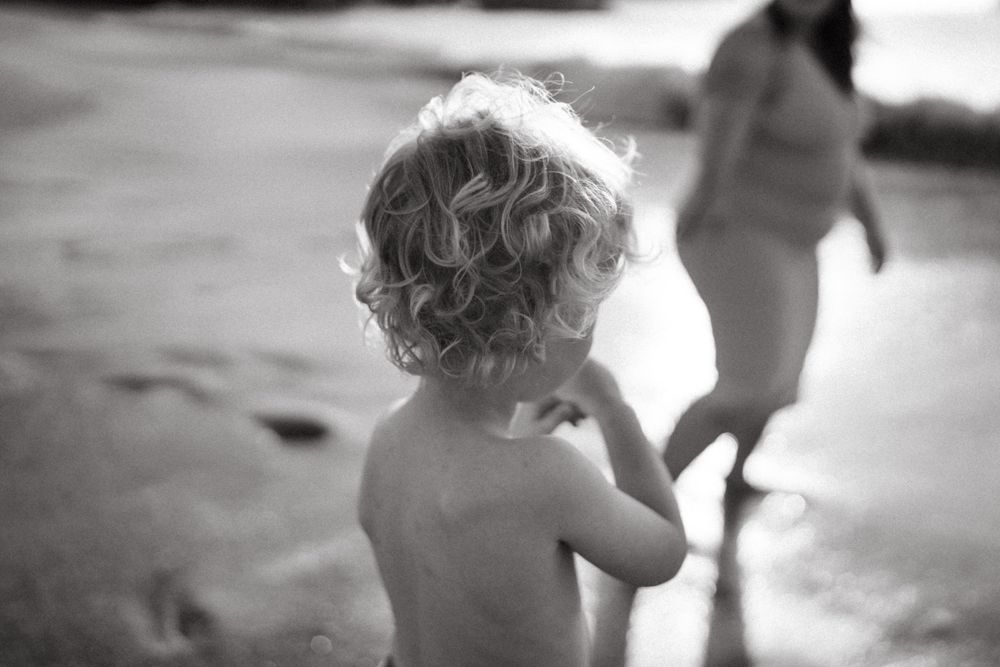 Toddler with curly hair watching figure walking on beach in black and white.
