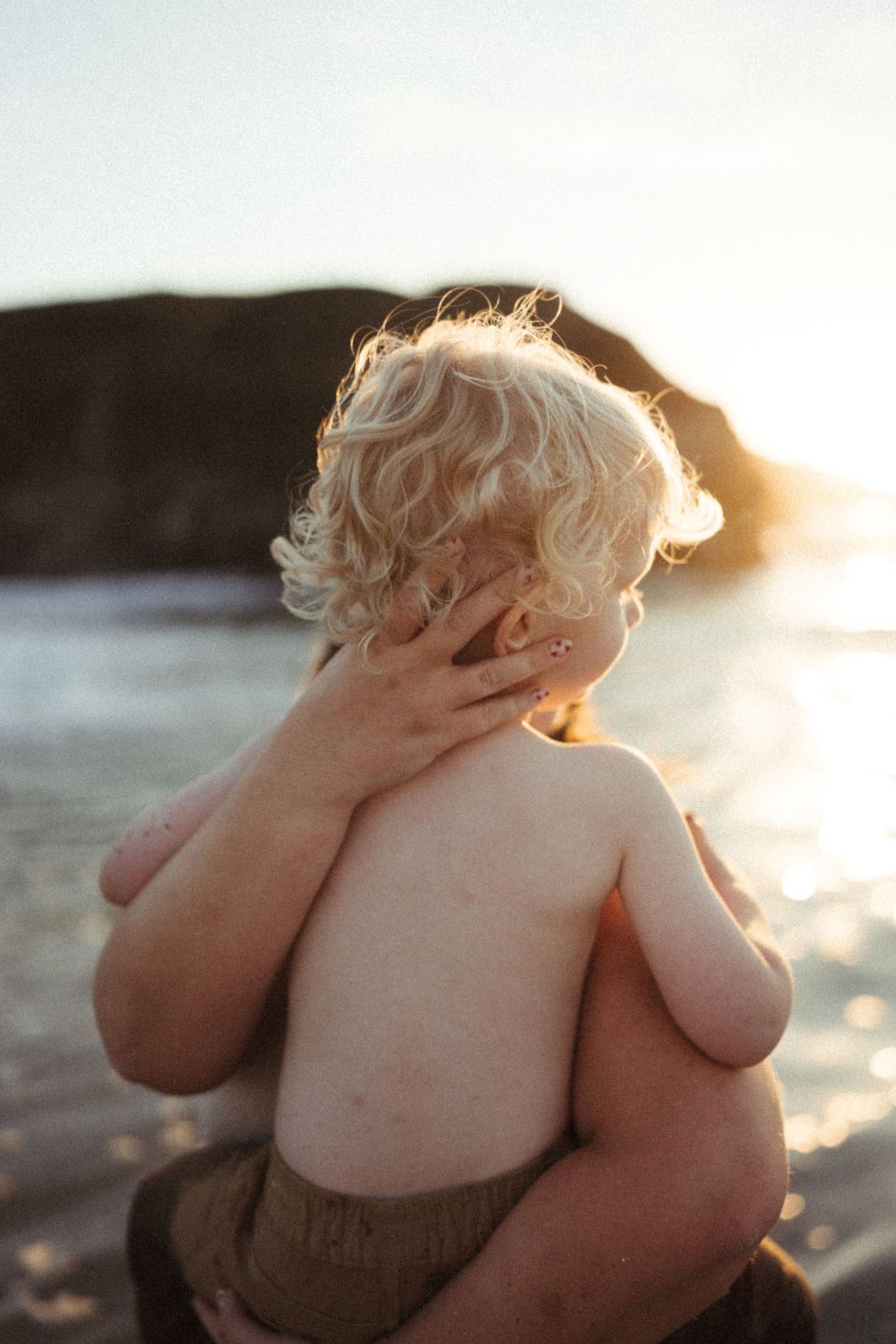 Small child with curly blonde hair covering face while sitting at beach during sunset.