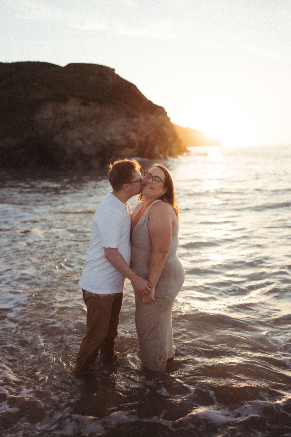Romantic couple embracing in ocean water at sunset with rocky cliffs in background.