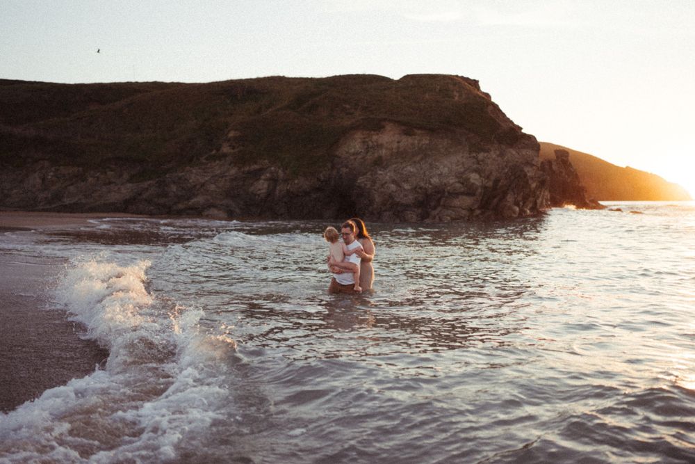 Couple playing in ocean waves at sunset near rocky coastline.