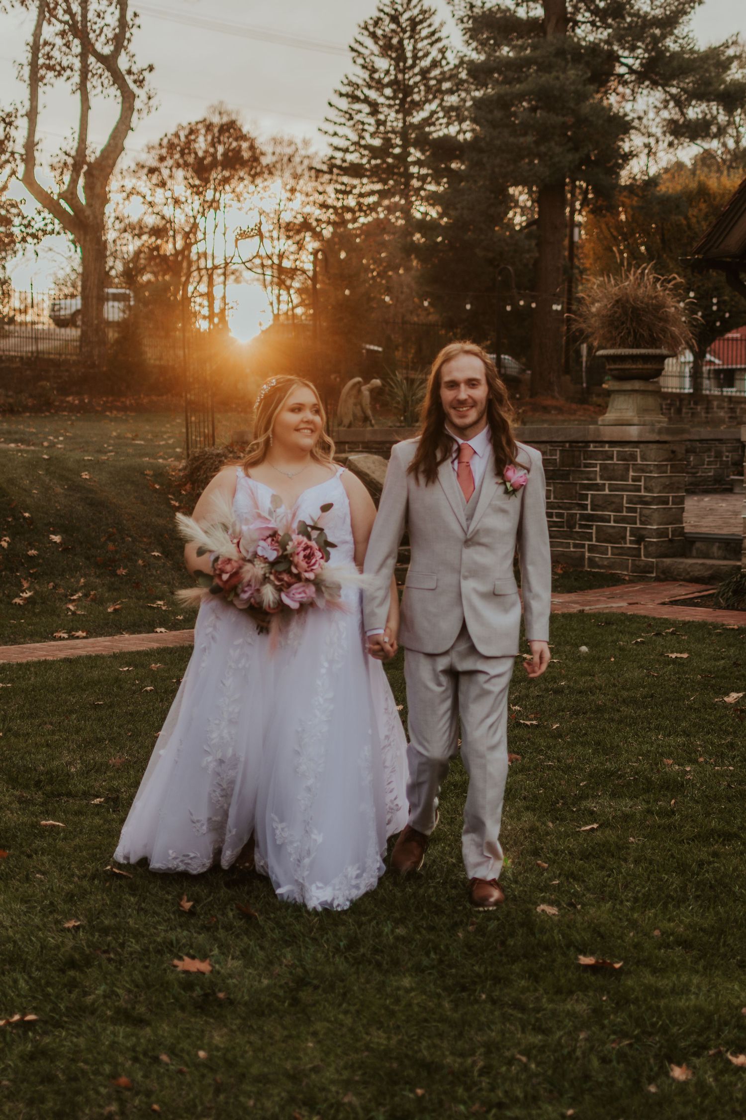 A couple walks hand in hand through a park at sunset after their wedding ceremony.