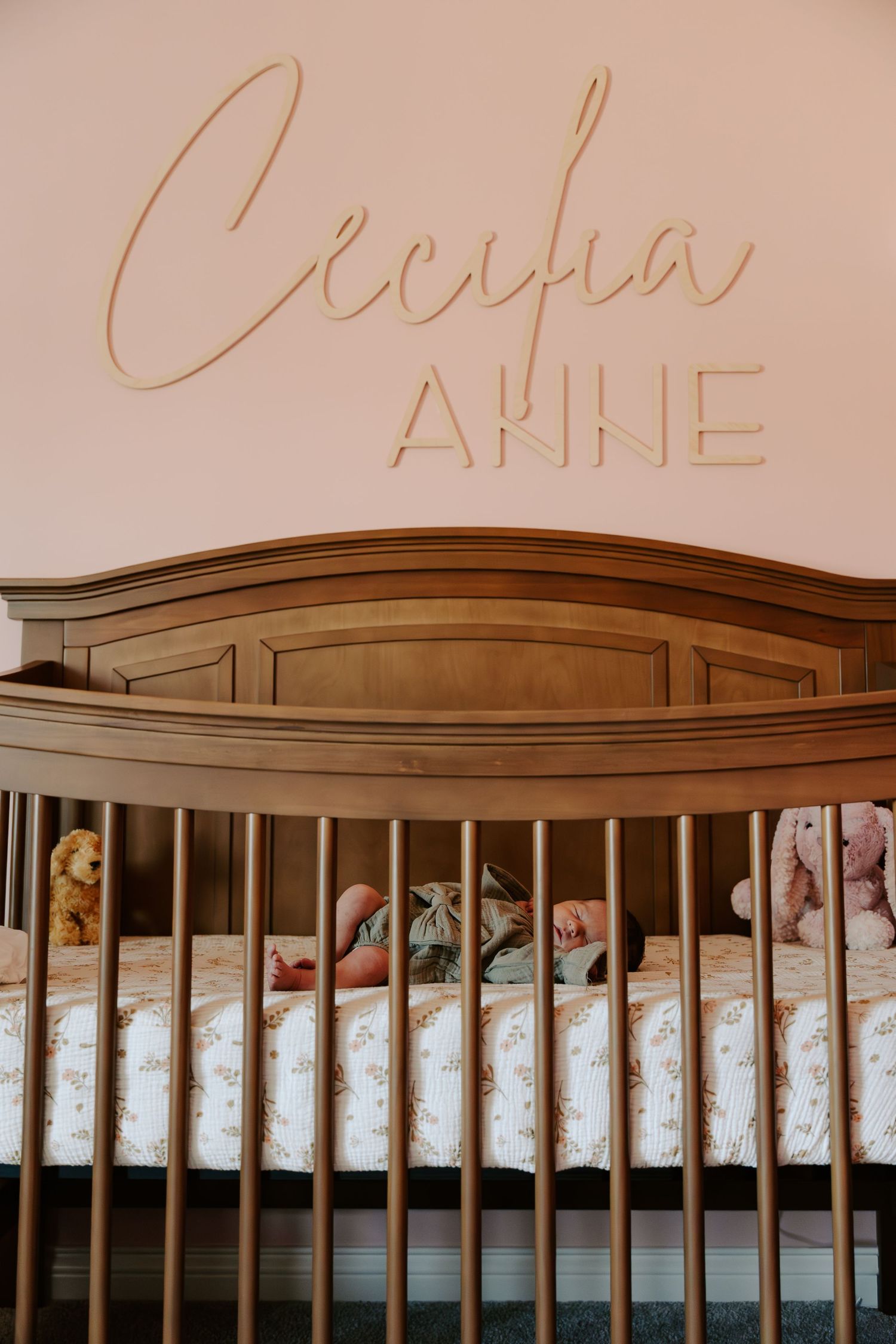 Wooden baby crib with curved headboard and name 'Anne' written above in cursive lettering against pink wall.