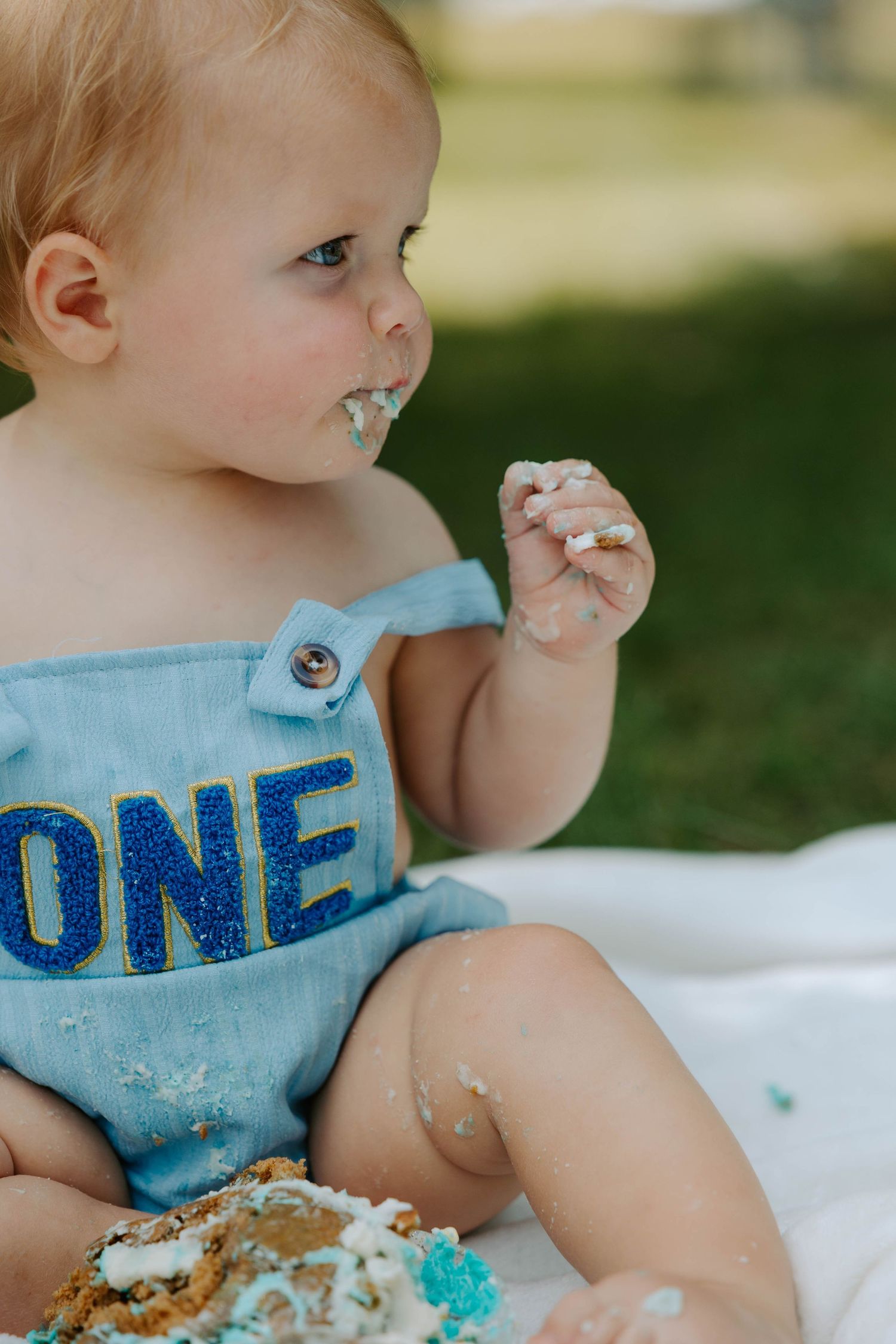 A baby wearing a light blue bib with 'ONE' text smashes and enjoys their first birthday cake with blue frosting.