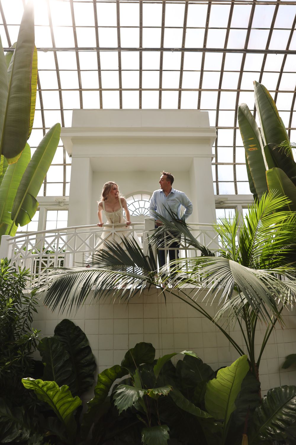 Wedding couple standing on balcony in conservatory surrounded by tropical plants and natural light.