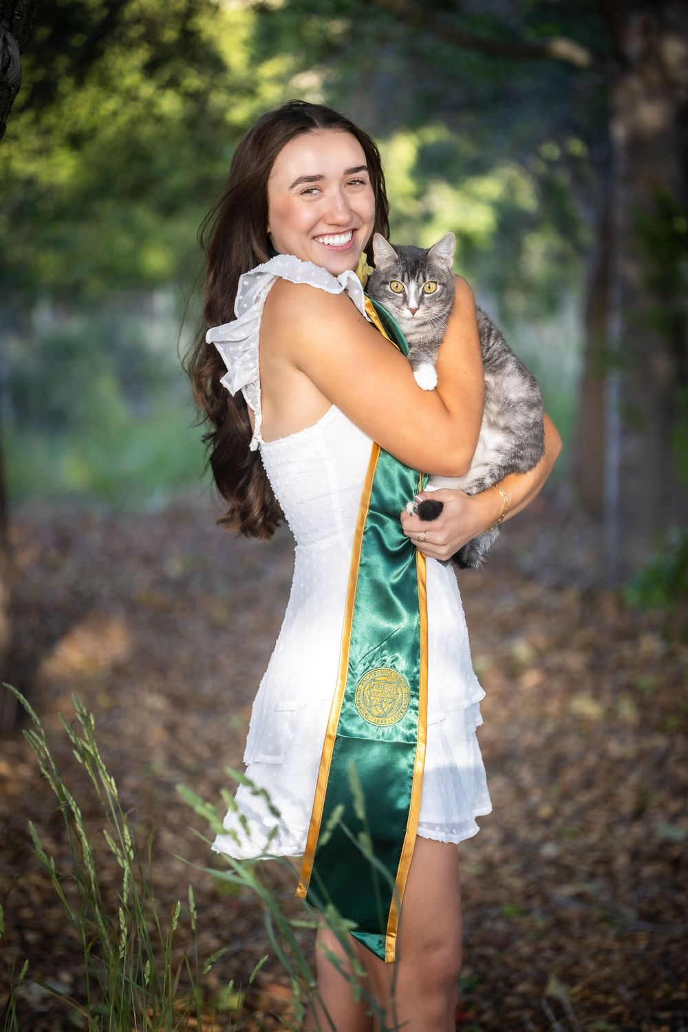 A series of outdoor photos showing someone in a white dress and green sash holding and cuddling a gray tabby cat in nature.