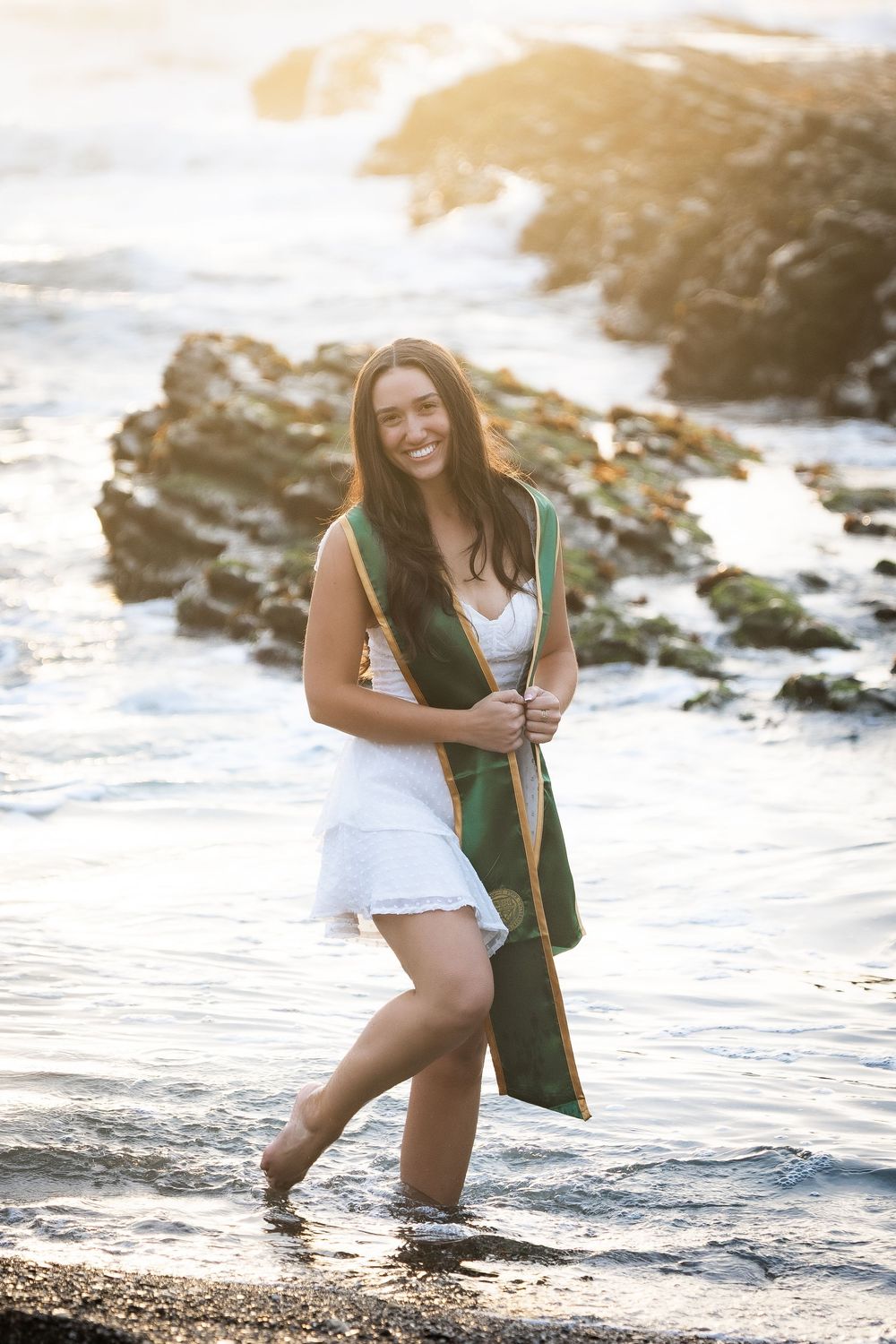 A woman in a white swimsuit poses playfully in shallow ocean water at sunset.