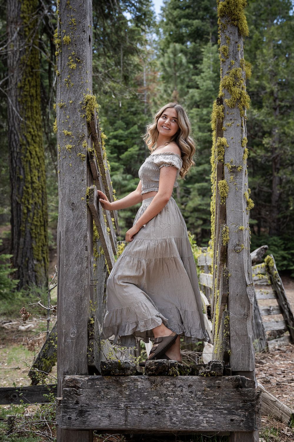 A person in a flowy beige dress poses gracefully among mossy tree trunks in a serene forest setting.