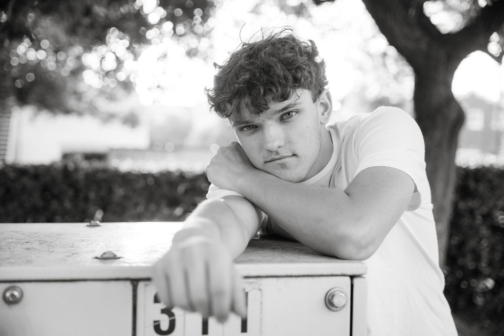 Black and white portrait of someone leaning on a white surface outdoors with trees in the background.