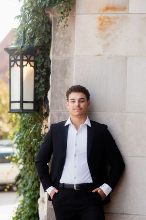 A person in a black suit and white shirt poses against a stone wall with ivy and a decorative lantern.