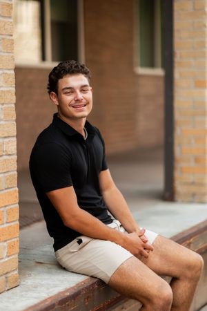 A young person in a black polo shirt and khaki shorts sits casually on brick steps with a warm smile.