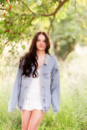Young woman with long dark hair wearing a white dress and oversized denim jacket standing outdoors near a cherry tree.