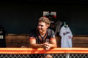 A person leans on an orange railing in a baseball dugout with uniforms visible in the background.