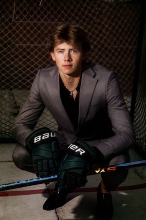 A hockey player in a grey suit poses with equipment against a dark background for a professional portrait series.