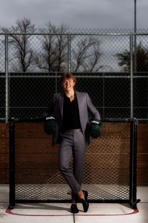 A person in a suit leans against a chain link fence at a baseball field during dusk.