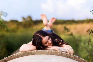Young woman with dark hair and tattoos resting on a concrete ledge outdoors in a lush green field.