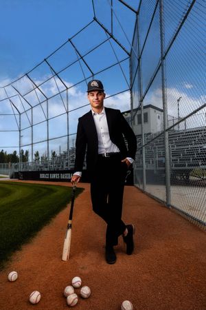 A figure in formal attire with a baseball bat stands on a baseball field near scattered baseballs and batting cage netting.