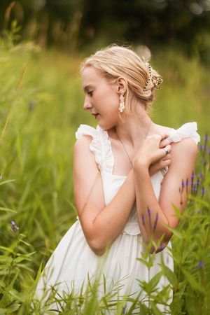 A person in a white dress sits thoughtfully in a summer meadow with wildflowers and tall grass.